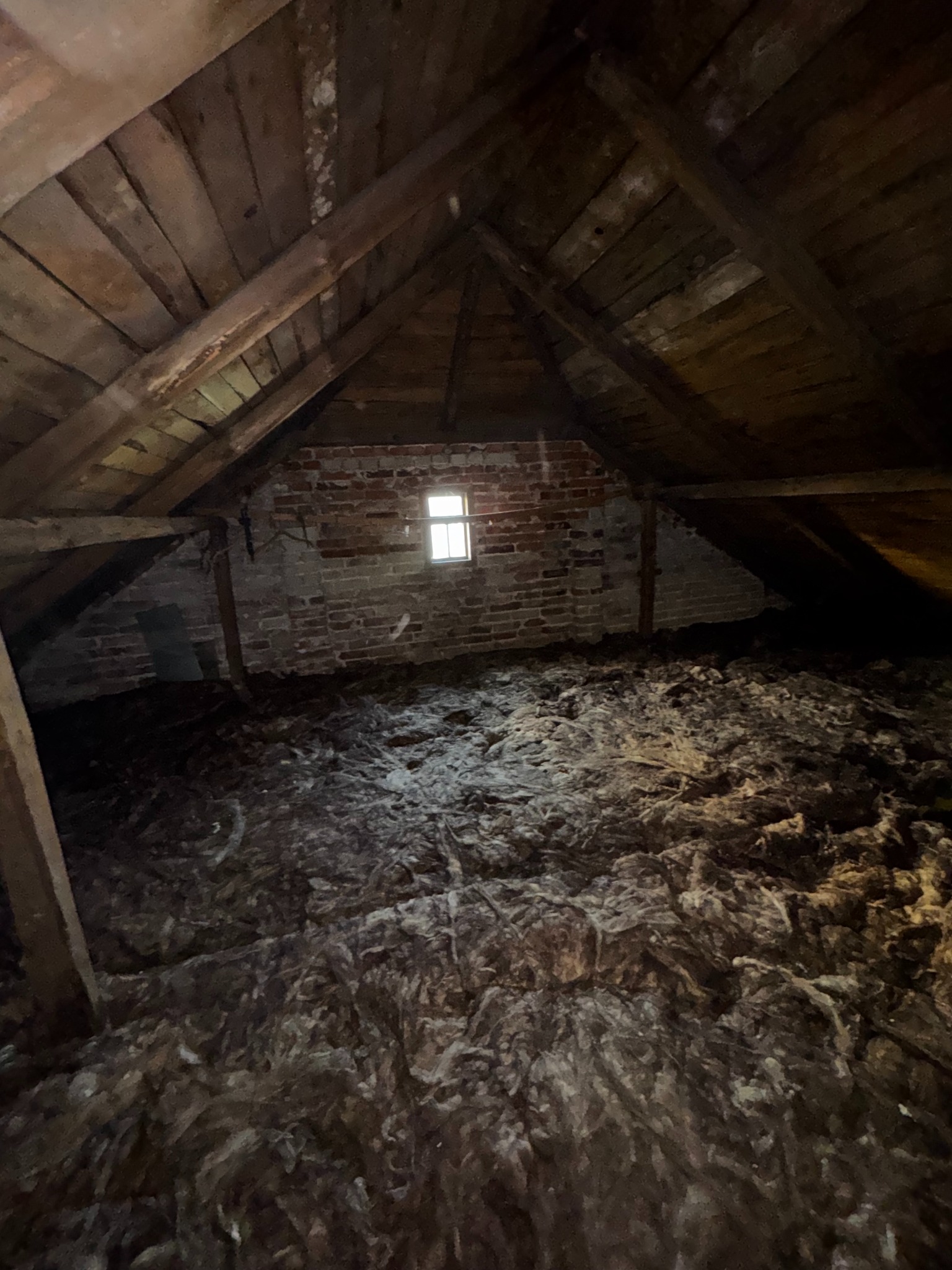 Poddasze z drewnianą konstrukcją i oknem w ceglanej ścianie, wypełnione materiałem izolacyjnym. Widok z góry na rozłożoną warstwę ocieplenia.