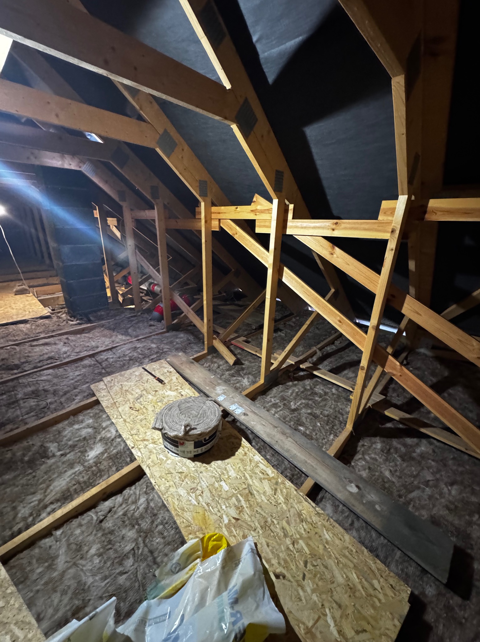 Poddasze w trakcie prac izolacyjnych: wełna mineralna między krokwiami, widoczne elementy konstrukcji drewnianej i rolka sznurka. Ujęcie z dołu, ciemne tło.