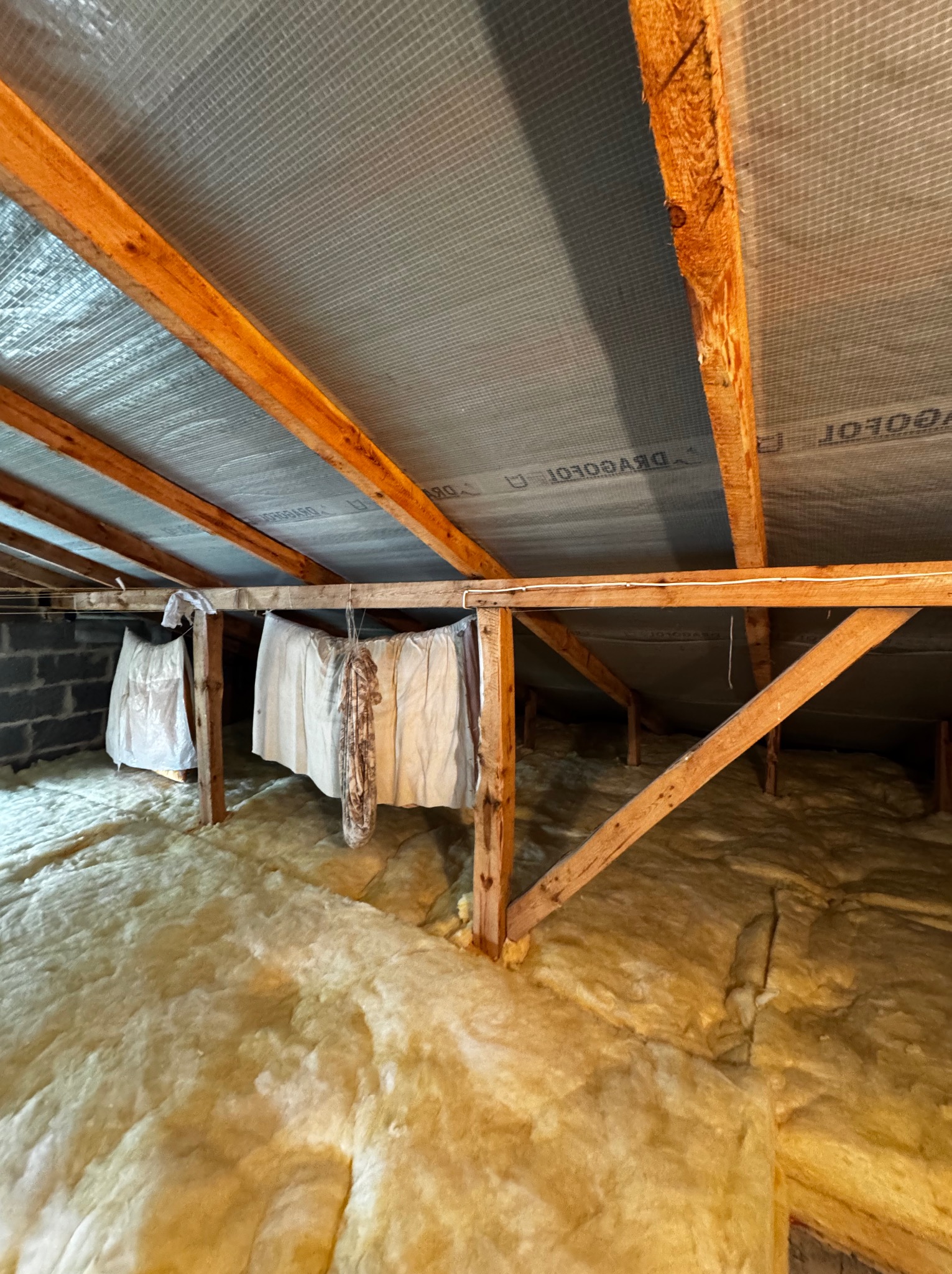 Poddasze z widoczną izolacją z wełny mineralnej, drewnianą konstrukcją więźby dachowej i folią paroizolacyjną. Część poddasza zasłonięta białym materiałem.