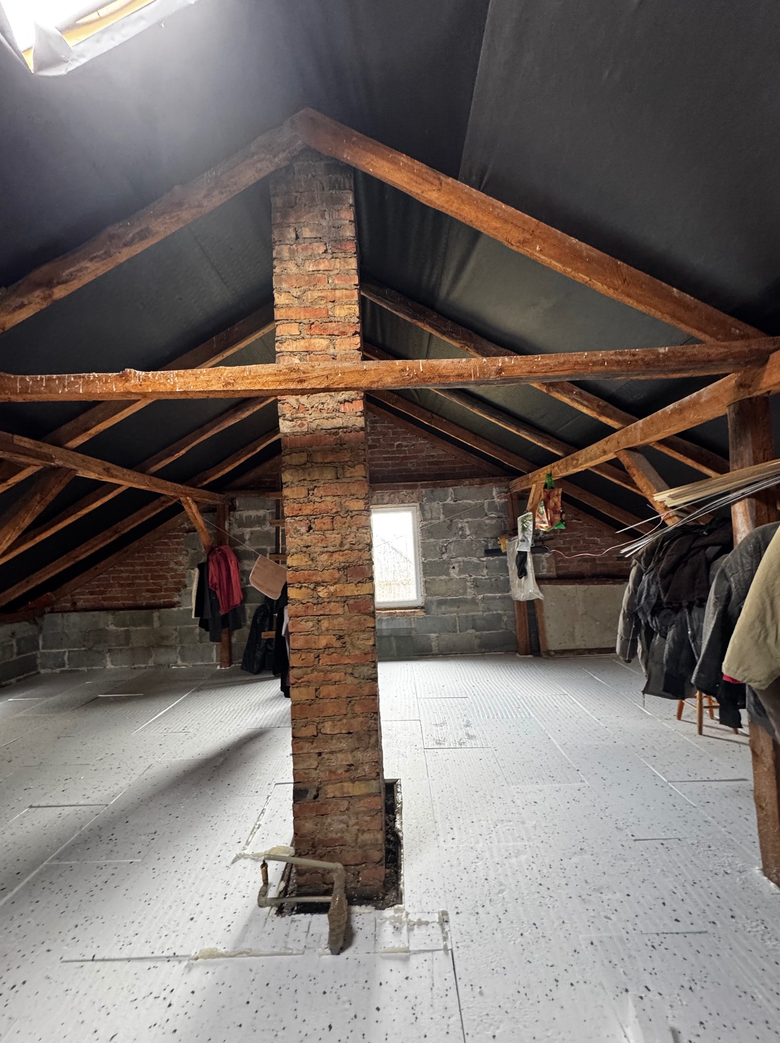 Poddasze z widoczną konstrukcją drewnianą i kominem z cegły, przygotowane do ocieplenia w Skierniewicach. Na podłodze białe płyty izolacyjne.