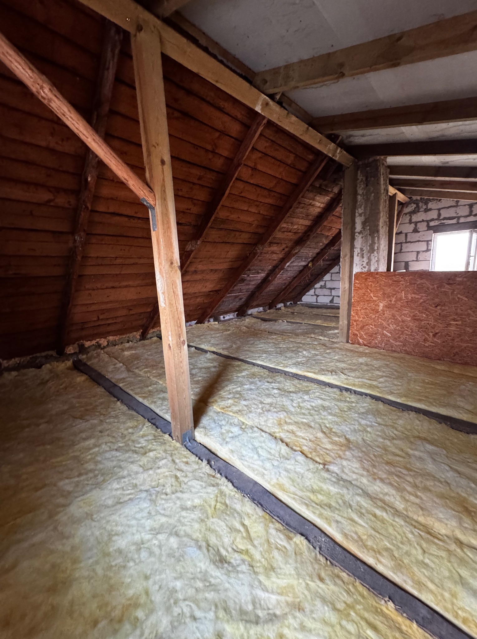 Poddasze z drewnianą konstrukcją i izolacją z wełny mineralnej, widoczne elementy muru i okno. Izolacja ułożona między legarami.