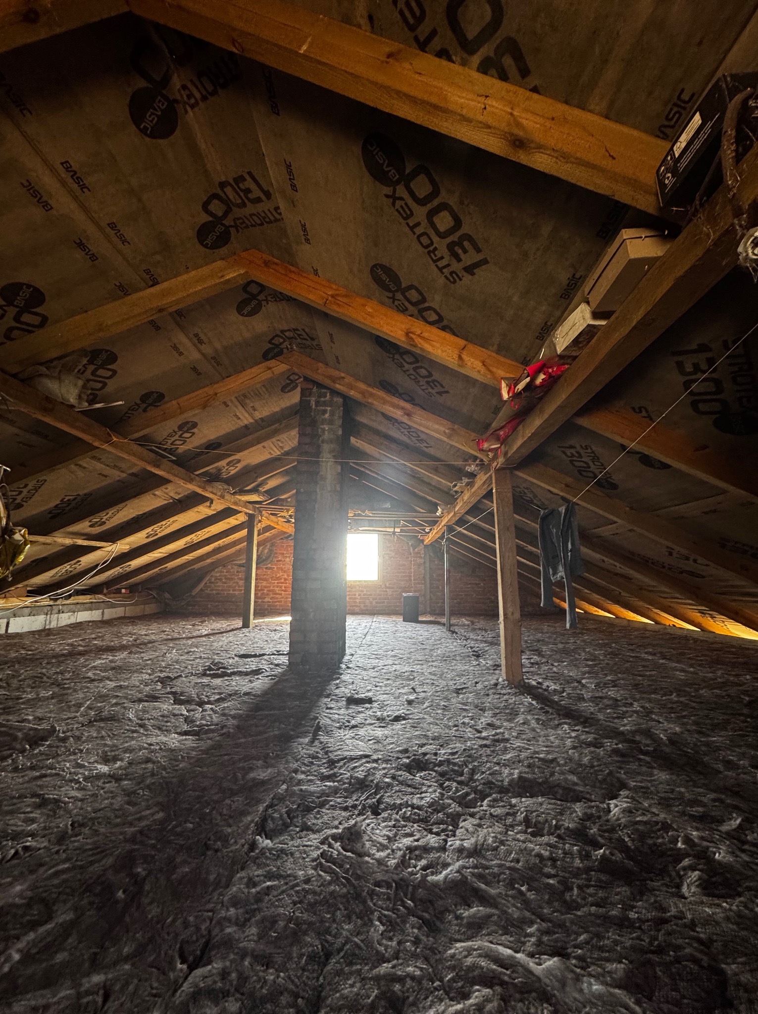 Poddasze po ociepleniu wełną mineralną, widoczne krokwie i membrana Strotex Basic 1300. W tle komin z cegły i okno w szczycie. Uwagę zwraca ubranie wiszące na sznurku.