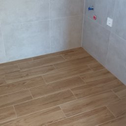 Paweł Lenarczyk - Wykończona łazienka z beżowymi płytkami imitującymi drewno na podłodze i szarymi płytkami na ścianach. Widoczne przyłącza wodne i gniazdko elektryczne.