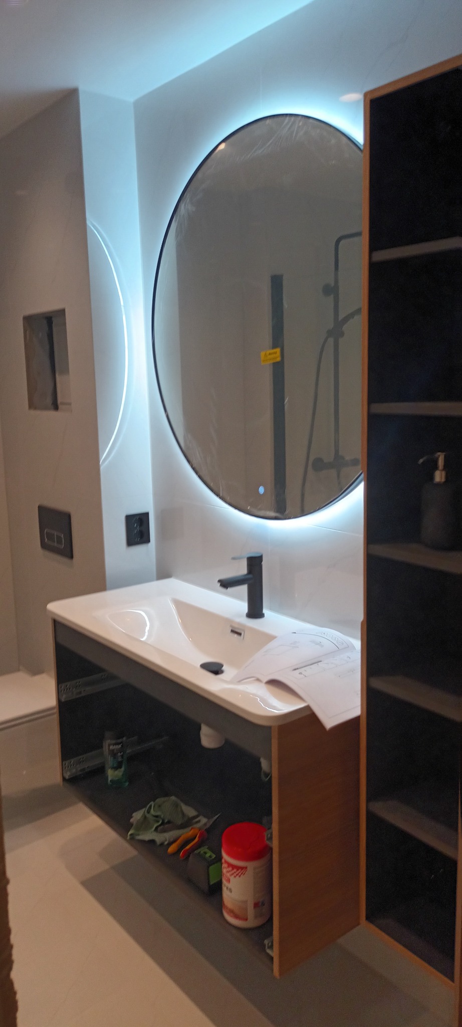 Nowoczesna łazienka z okrągłym lustrem LED, białą umywalką i drewnianą szafką. Widoczne narzędzia i akcesoria, sugerujące etap wykończeniowy.