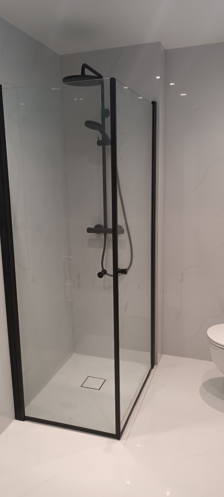 Nowoczesna kabina prysznicowa z czarnymi profilami i deszczownicą, białe płytki imitujące marmur, minimalistyczny odpływ liniowy. Wykończenie łazienki w stylu skandynawskim.