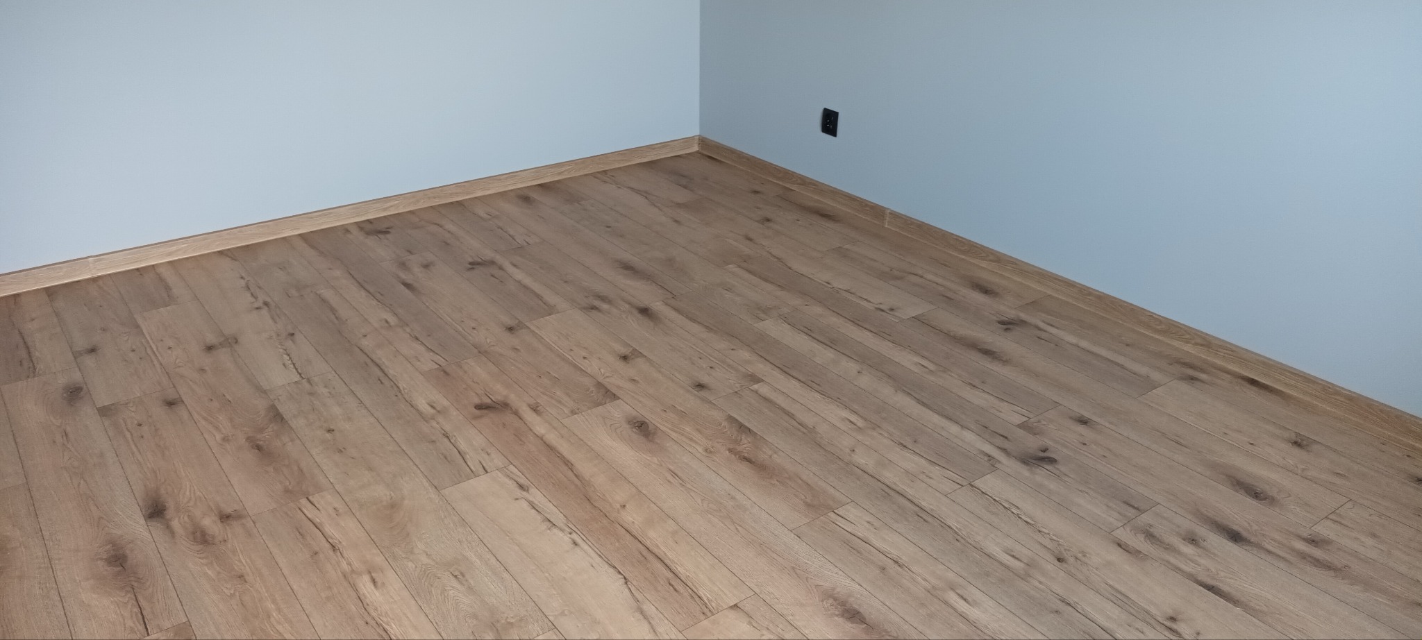 Ukończona podłoga z paneli w odcieniach brązu, z widoczną listwą przypodłogową i fragmentem ściany w kolorze jasnego błękitu z gniazdkiem elektrycznym.