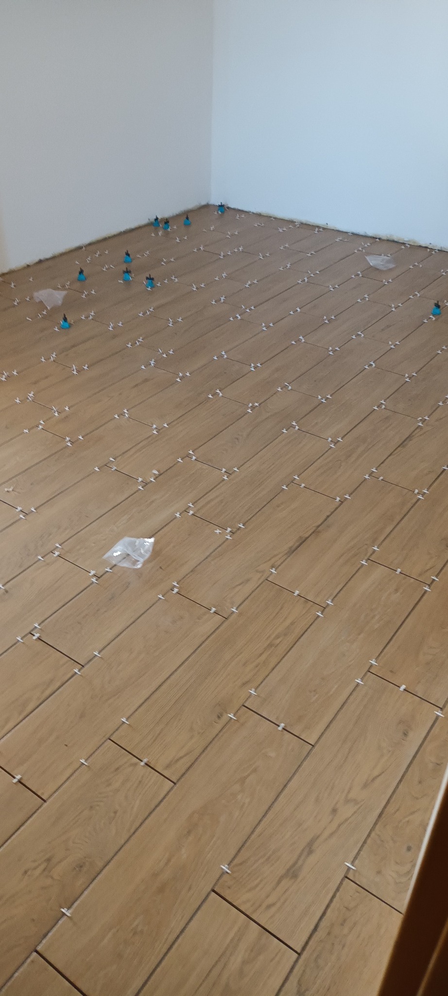 Ułożone płytki imitujące drewno w jasnym pomieszczeniu, z użyciem niebieskich klipsów poziomujących i białych krzyżyków dystansowych. Widok pod kątem.