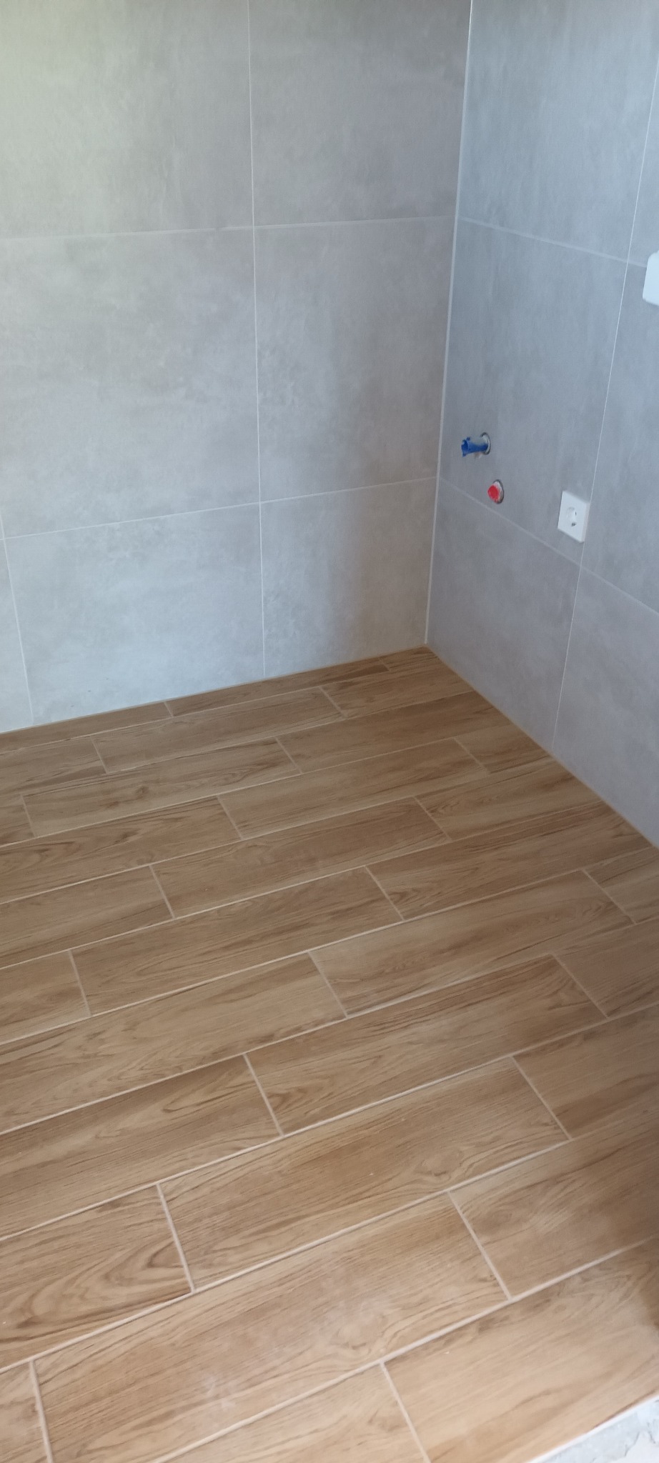 Wykończona łazienka z beżowymi płytkami imitującymi drewno na podłodze i szarymi płytkami na ścianach. Widoczne przyłącza wodne i gniazdko elektryczne.