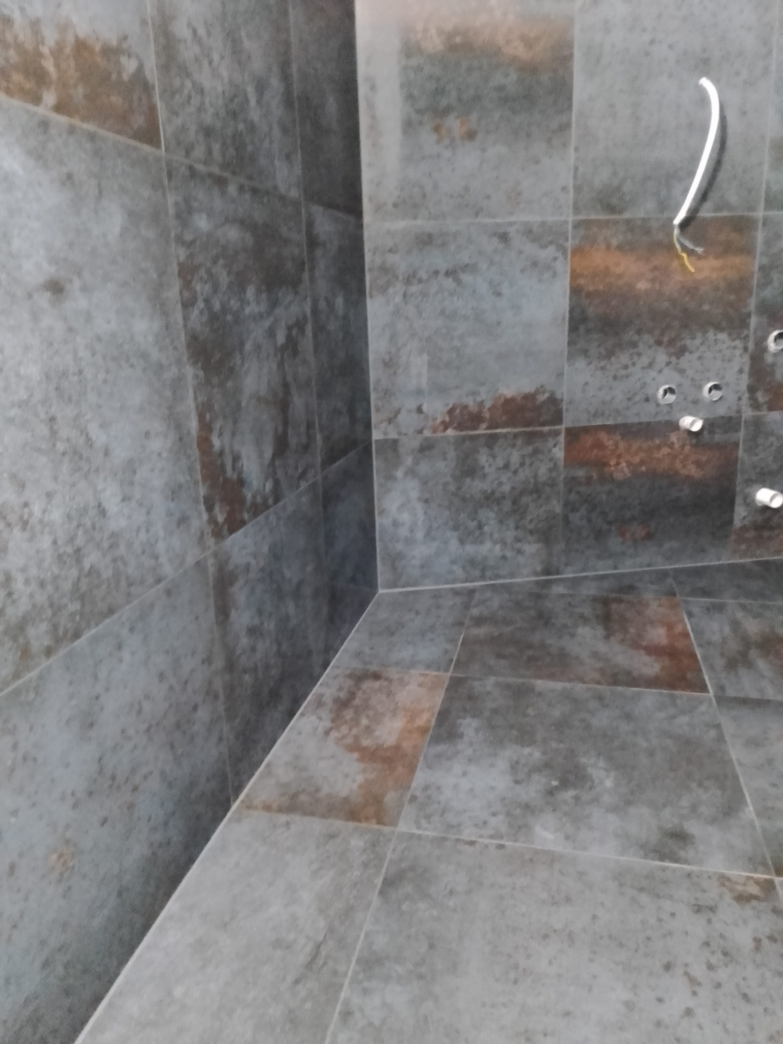 Wykończona łazienka z szarymi, matowymi płytkami imitującymi kamień. Widoczne otwory na armaturę i wystający kabel. Ujęcie z narożnika pomieszczenia.