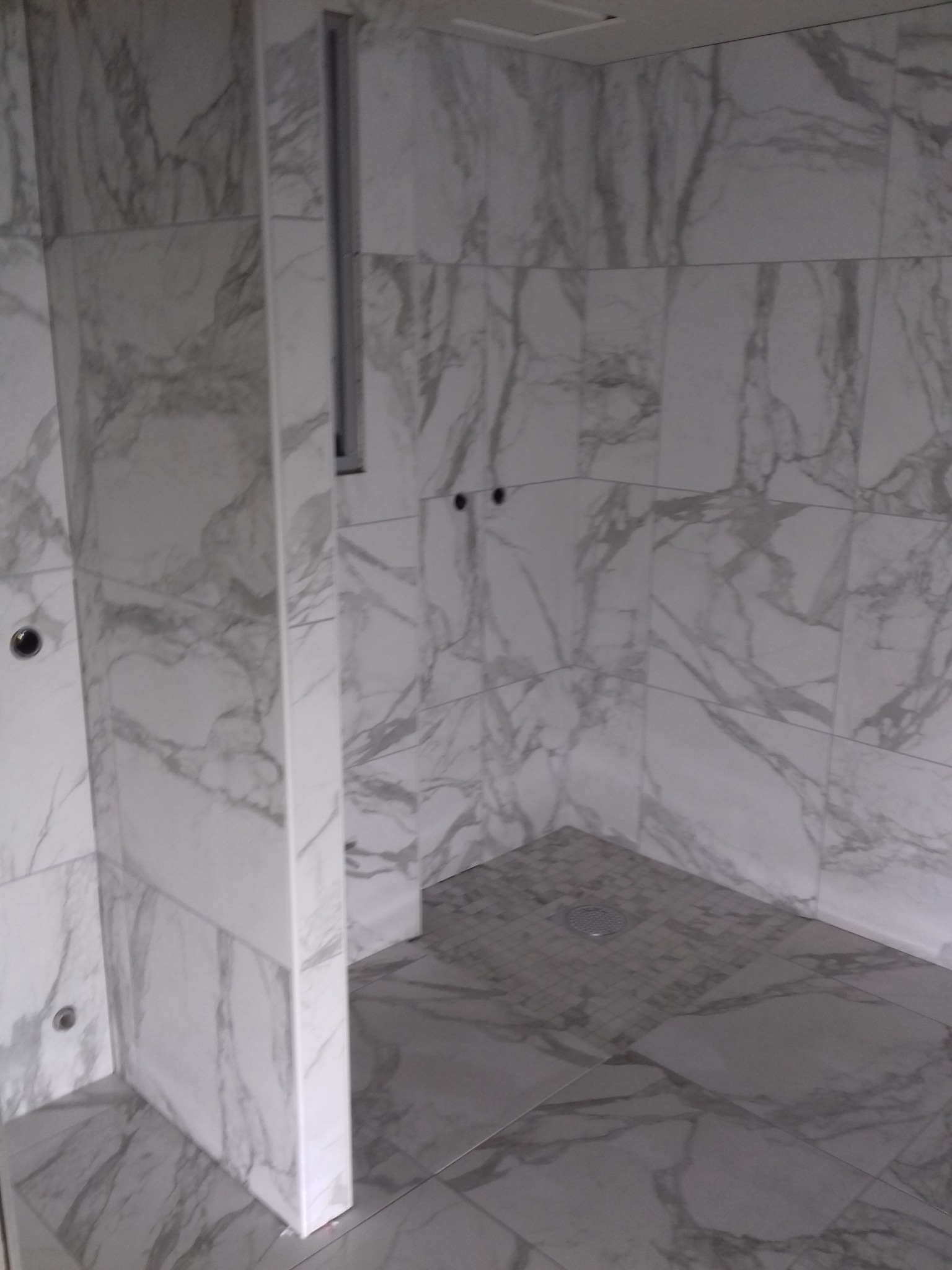 Wykończona kabina prysznicowa z szarymi płytkami imitującymi marmur na ścianach i podłodze. Widoczne odpływ liniowy i otwory na armaturę.