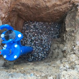 Przedsiębiorstwo Handlowo usługowe Kapi-Kop Dominik Olszak - Niebieski zawór przyłączeniowy w wykopie wypełnionym żwirem, widok z góry. Wykop w ziemi o nierównych ścianach.