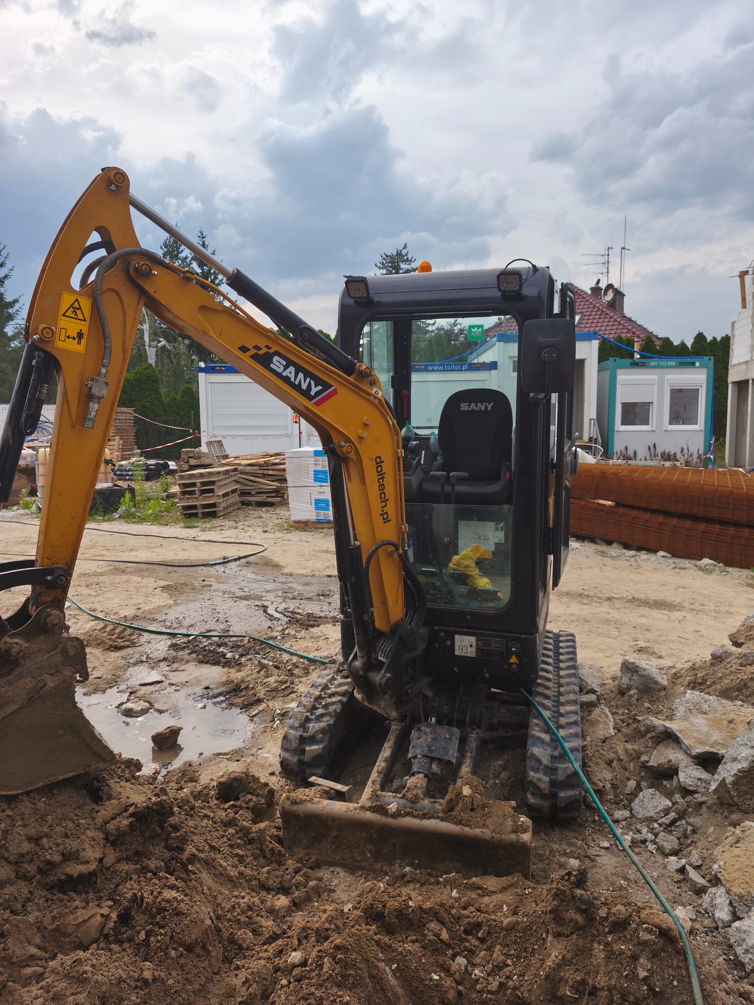 Żółto-czarna mini koparka SANY w trakcie prac ziemnych na placu budowy, widoczne materiały budowlane i zbrojenia. Pochmurne niebo w tle.