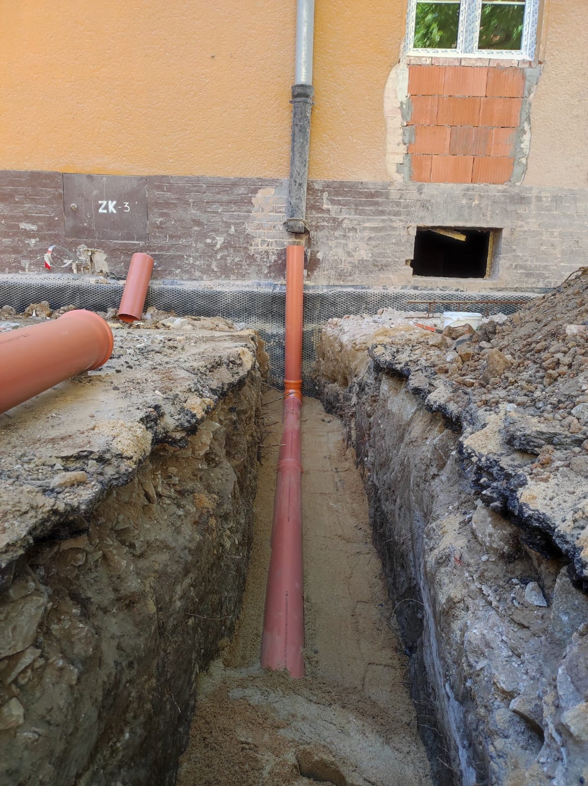 Wykop z ułożonym rurociągiem kanalizacyjnym z pomarańczowych rur. Widoczny fragment budynku z oknem i otworem w fundamencie. Prace ziemne w toku.