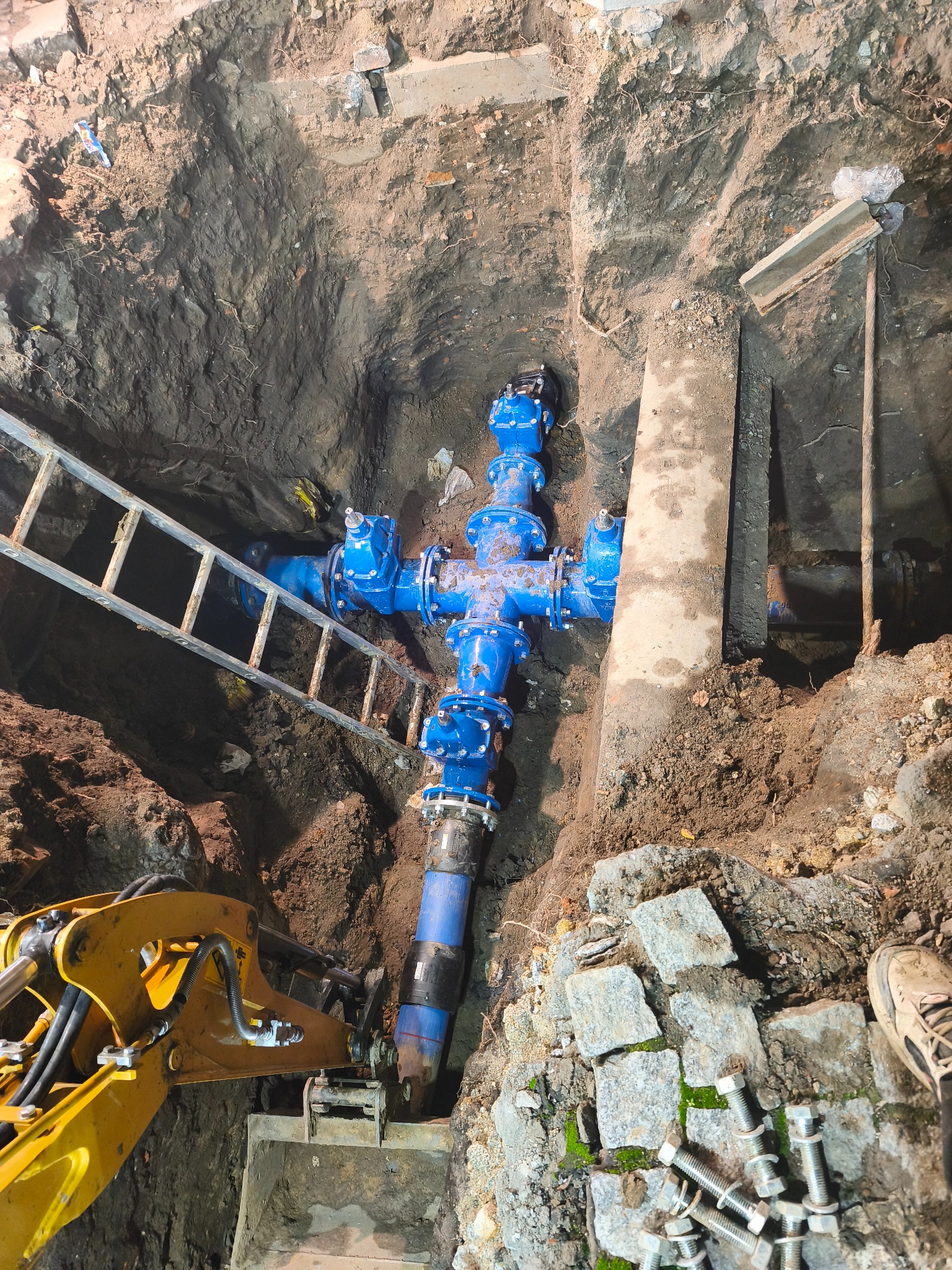 Niebieskie przyłącze wodne w wykopie, widok z góry. Żółta koparka i drabina w tle. Prace instalacyjne we Wrocławiu.