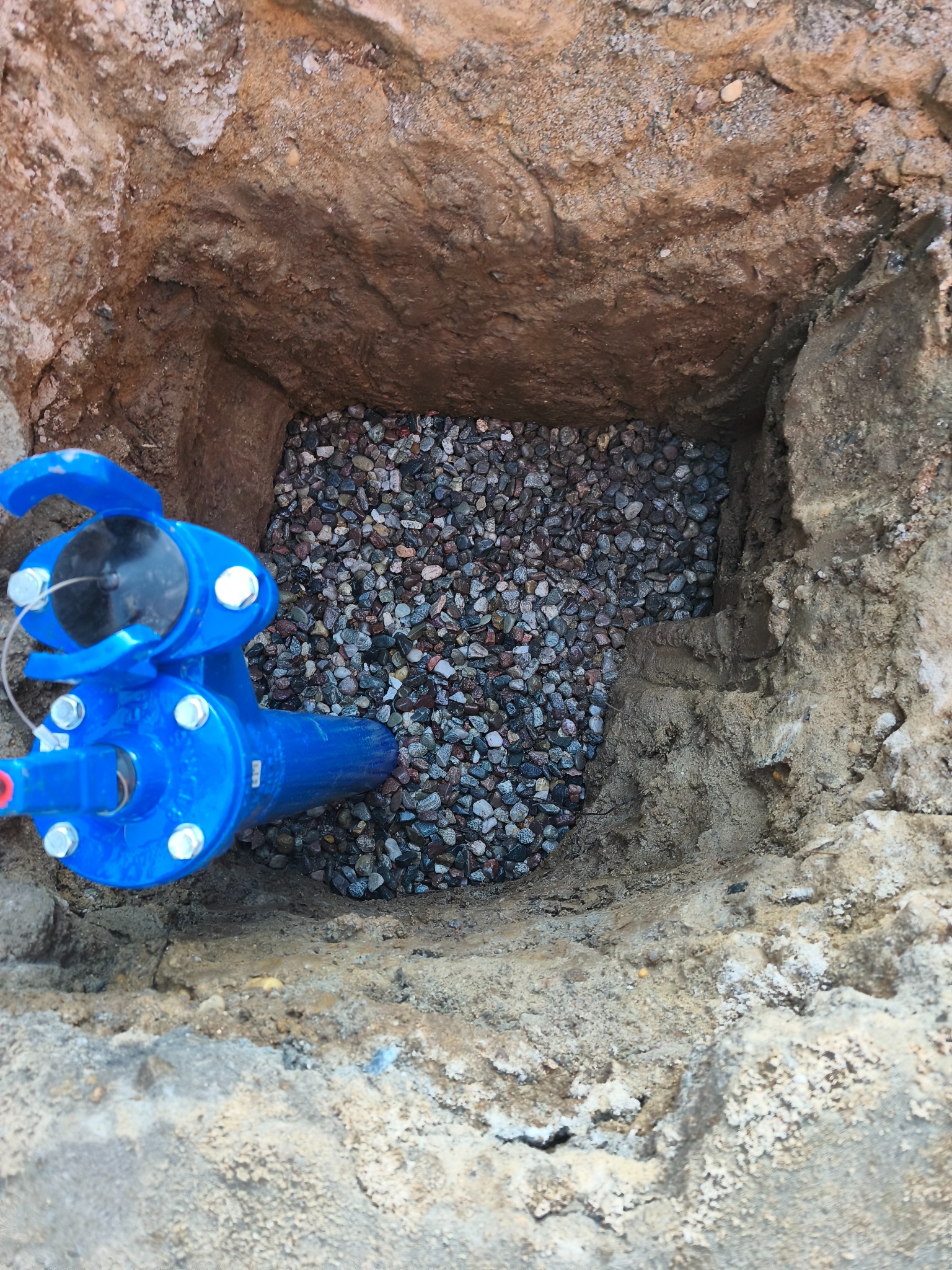 Niebieski zawór przyłączeniowy w wykopie wypełnionym żwirem, widok z góry. Wykop w ziemi o nierównych ścianach.