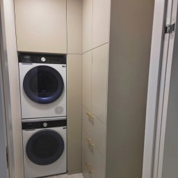 MEBLE ADOZ JAKUB PERZANOWSKI - Beżowa zabudowa z pralką i suszarką w kolumnie, złote uchwyty szuflad, marmurowa podłoga. Funkcjonalne rozwiązanie do małej łazienki.