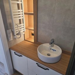 MEBLE ADOZ JAKUB PERZANOWSKI - Nowoczesna łazienka z białą szafką pod umywalkę z drewnianym blatem i czarnymi uchwytami. Nad szafką grzejnik drabinkowy i oświetlona półka w drewnianej obudowie.