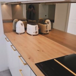 MEBLE ADOZ JAKUB PERZANOWSKI - Białe szafki kuchenne z drewnianym blatem i złotymi uchwytami. Na blacie czarna płyta indukcyjna i sprzęt AGD marki SMEG. Lustrzane wykończenie górnych szafek.