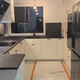 MEBLE ADOZ JAKUB PERZANOWSKI - Jasna kuchnia z czarnymi akcentami: blat, armatura i płytki. Podłoga przygotowana do położenia płytek, wyznaczona pomarańczową taśmą. Widok na okno i oświetlenie.