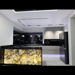 MEBLE ADOZ JAKUB PERZANOWSKI - Nowoczesna kuchnia z wyspą oświetloną od dołu, fronty w odcieniach szarości i czerni, minimalistyczne oświetlenie sufitowe. Blat wyspy z czarnego kamienia.