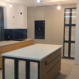 Nowoczesna kuchnia z wyspą i ryflowanymi szybami w zabudowie. Beżowe fronty szafek, czarny sprzęt AGD. Widoczne niedokończone prace wykończeniowe.