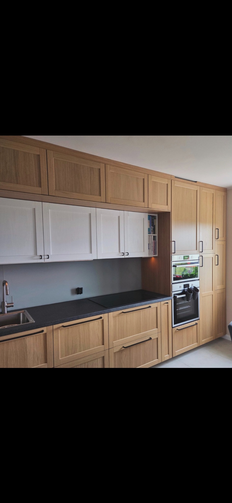 Jasna kuchnia z drewnianymi frontami i czarnym blatem. Górne szafki białe, nad blatem gniazdko elektryczne. Wbudowany piekarnik i szafka z książkami.