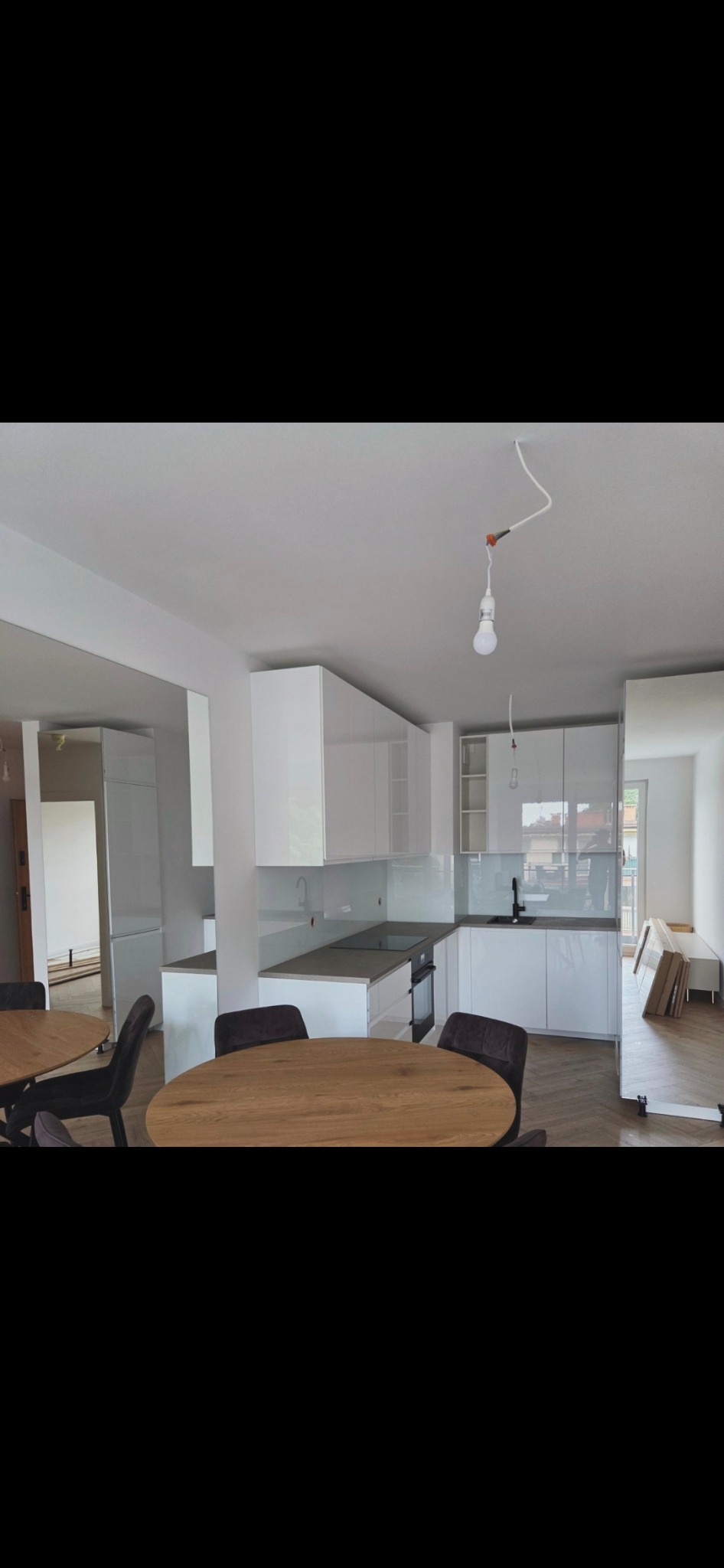 Jasna kuchnia z białymi szafkami i szarym blatem, okrągłe stoły z ciemnymi krzesłami. Widoczne niedokończone prace, wisząca żarówka bez klosza.