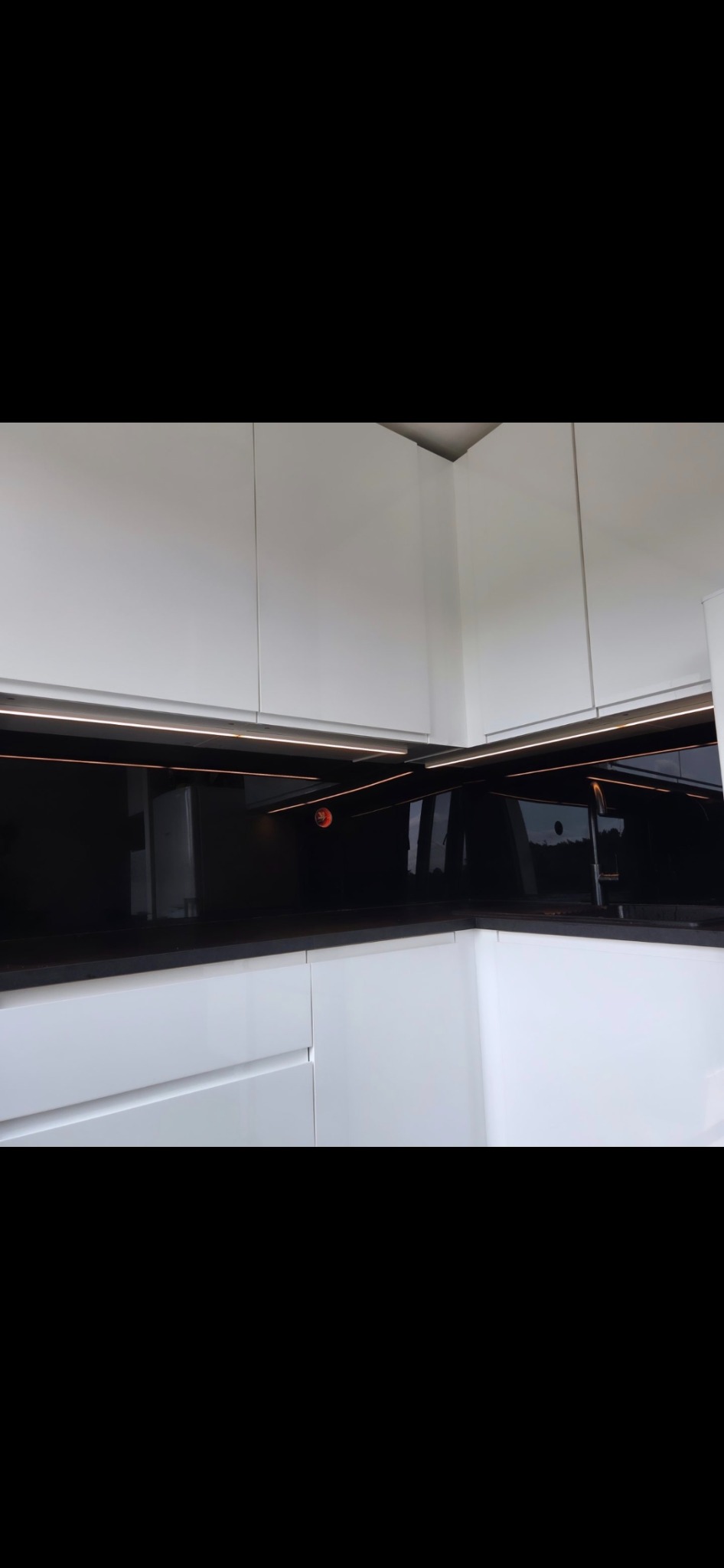 Biała, nowoczesna kuchnia z czarnym blatem i podświetleniem LED pod szafkami wiszącymi, ukazana w ujęciu z dołu, podkreślającym minimalistyczny design.