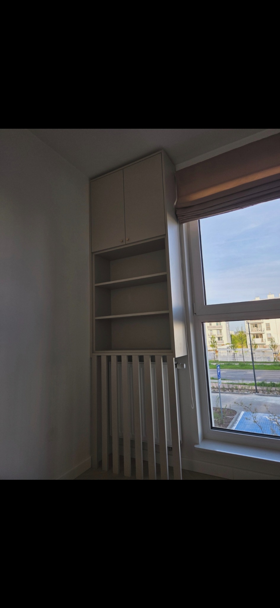 Biała szafa wnękowa z półkami nad grzejnikiem, widok z boku, jasne wnętrze, okno z roletą w tle, minimalistyczny design.