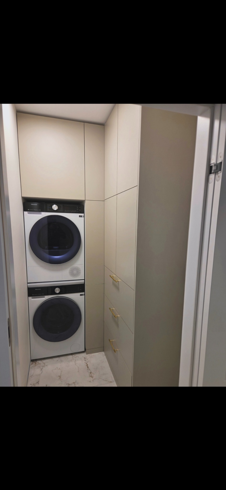 Beżowa zabudowa z pralką i suszarką w kolumnie, złote uchwyty szuflad, marmurowa podłoga. Funkcjonalne rozwiązanie do małej łazienki.