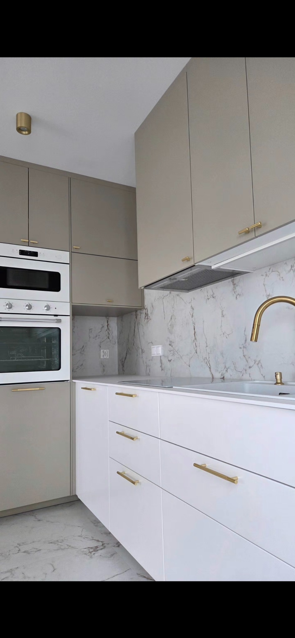 Nowoczesna kuchnia z białymi szafkami dolnymi i beżowymi górnymi, złote uchwyty, marmurowy backsplash i podłoga. Minimalistyczny design z piekarnikiem i płytą indukcyjną.