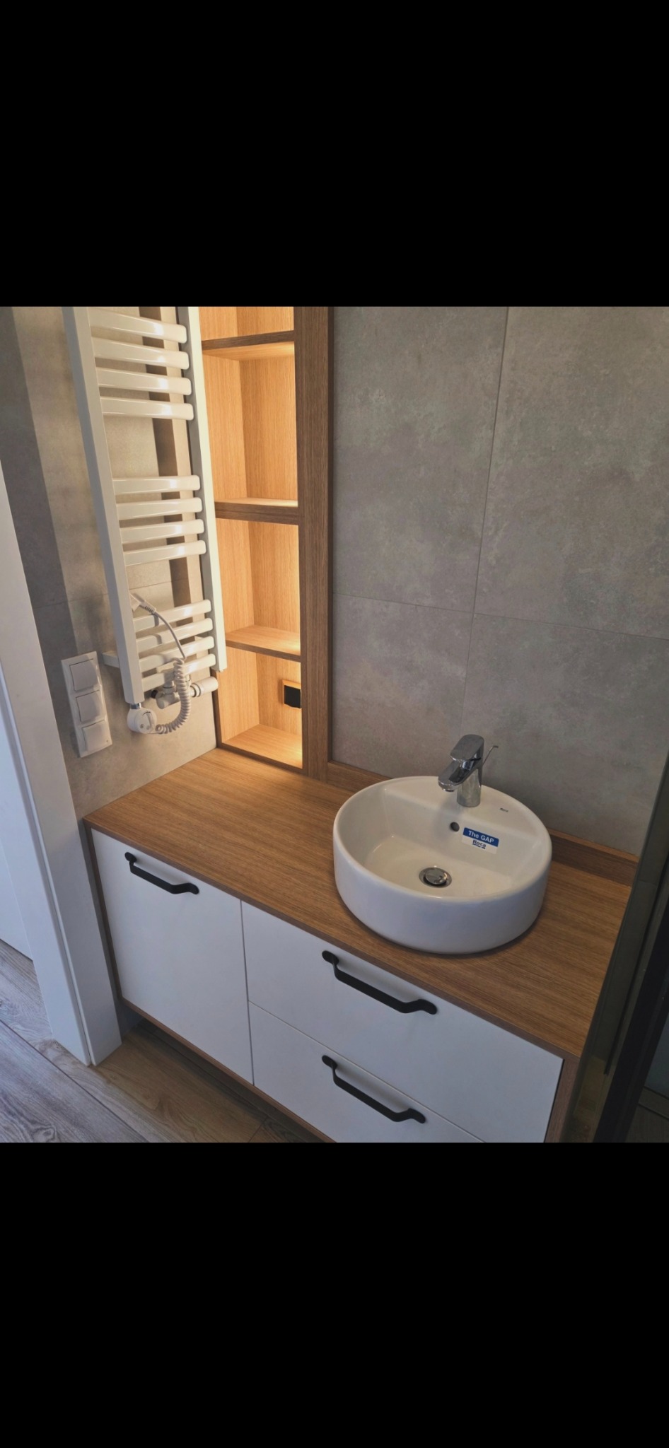 Nowoczesna łazienka z białą szafką pod umywalkę z drewnianym blatem i czarnymi uchwytami. Nad szafką grzejnik drabinkowy i oświetlona półka w drewnianej obudowie.