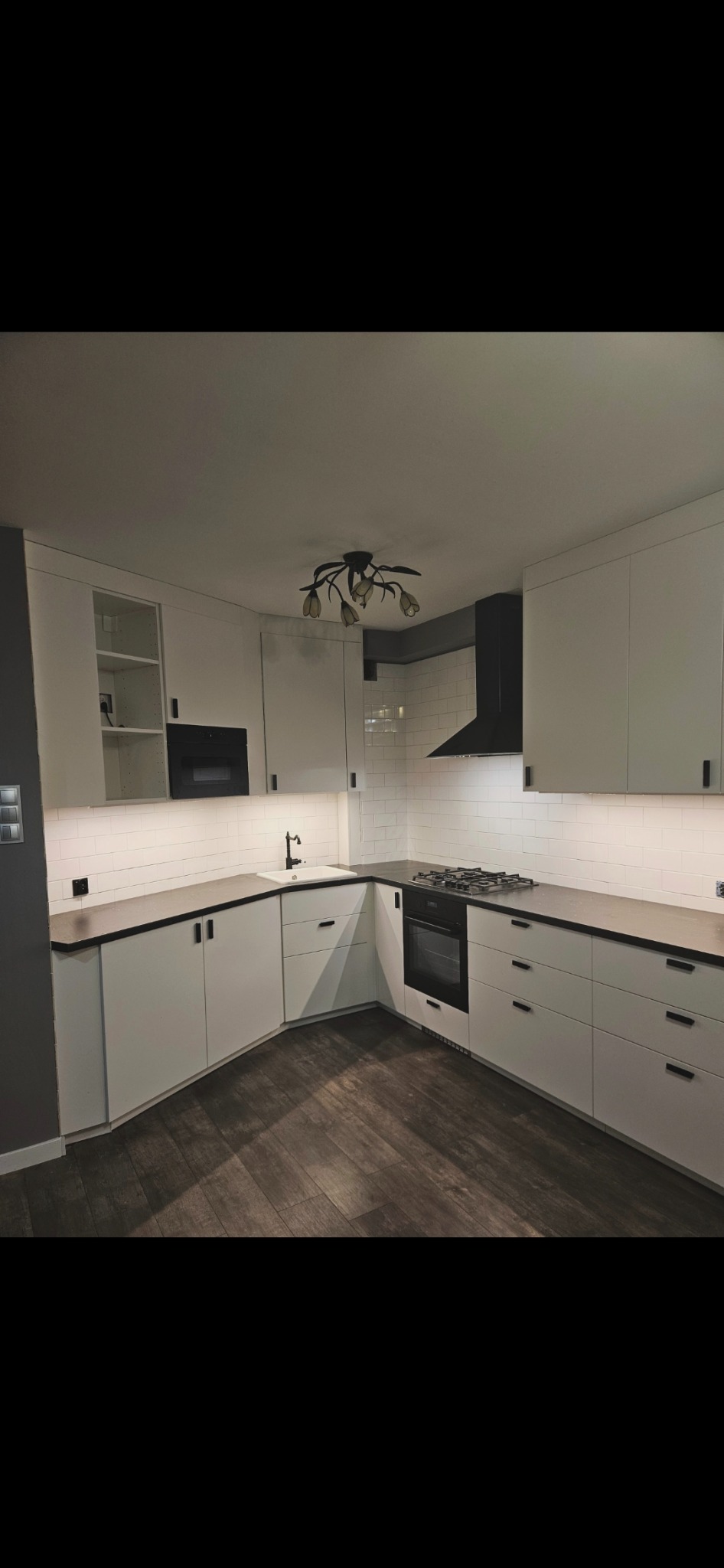 Nowoczesna kuchnia w bieli i czerni z drewnianą podłogą. Uwagę przykuwa designerska lampa sufitowa w kształcie kwiatów i czarny okap nad płytą gazową.