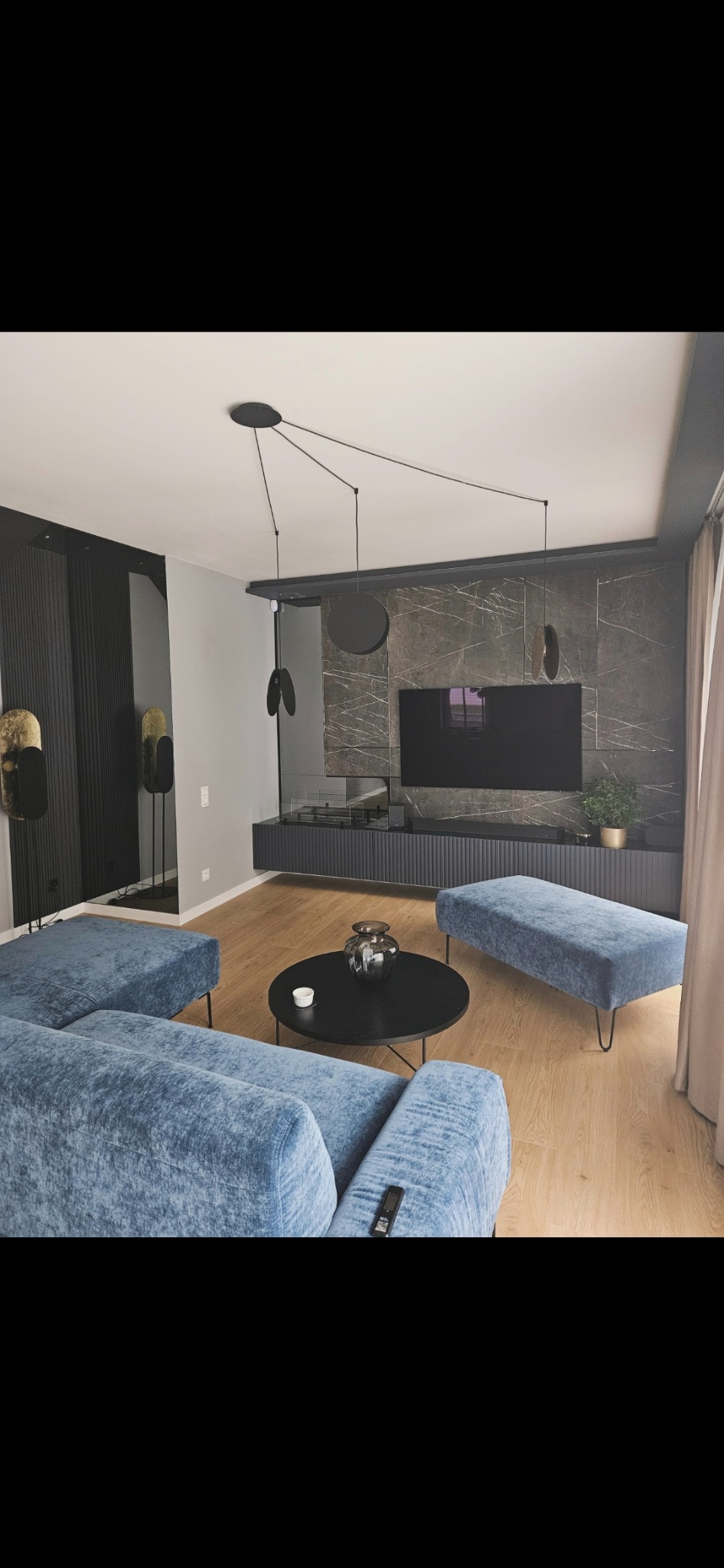 Nowoczesny salon z niebieską sofą, okrągłym stolikiem kawowym i ścianą TV z marmurowym wzorem. Minimalistyczna lampa wisząca i drewniana podłoga dodają elegancji.