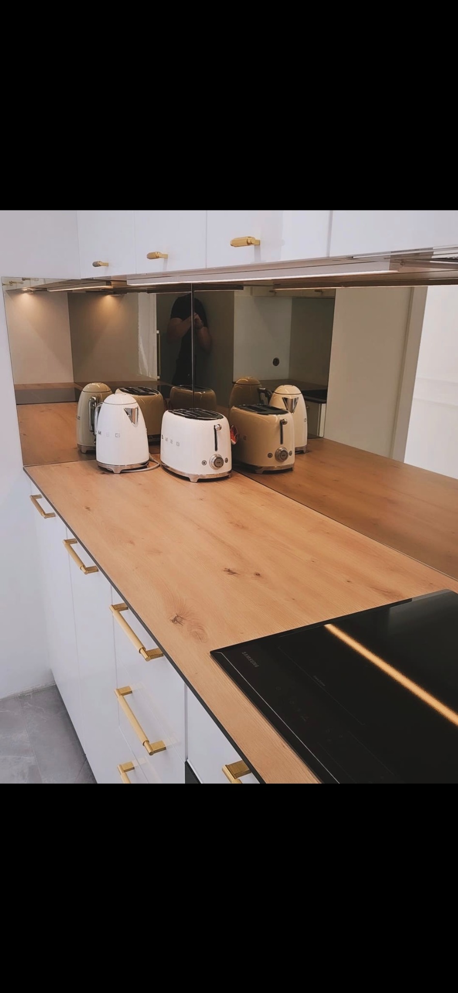 Białe szafki kuchenne z drewnianym blatem i złotymi uchwytami. Na blacie czarna płyta indukcyjna i sprzęt AGD marki SMEG. Lustrzane wykończenie górnych szafek.