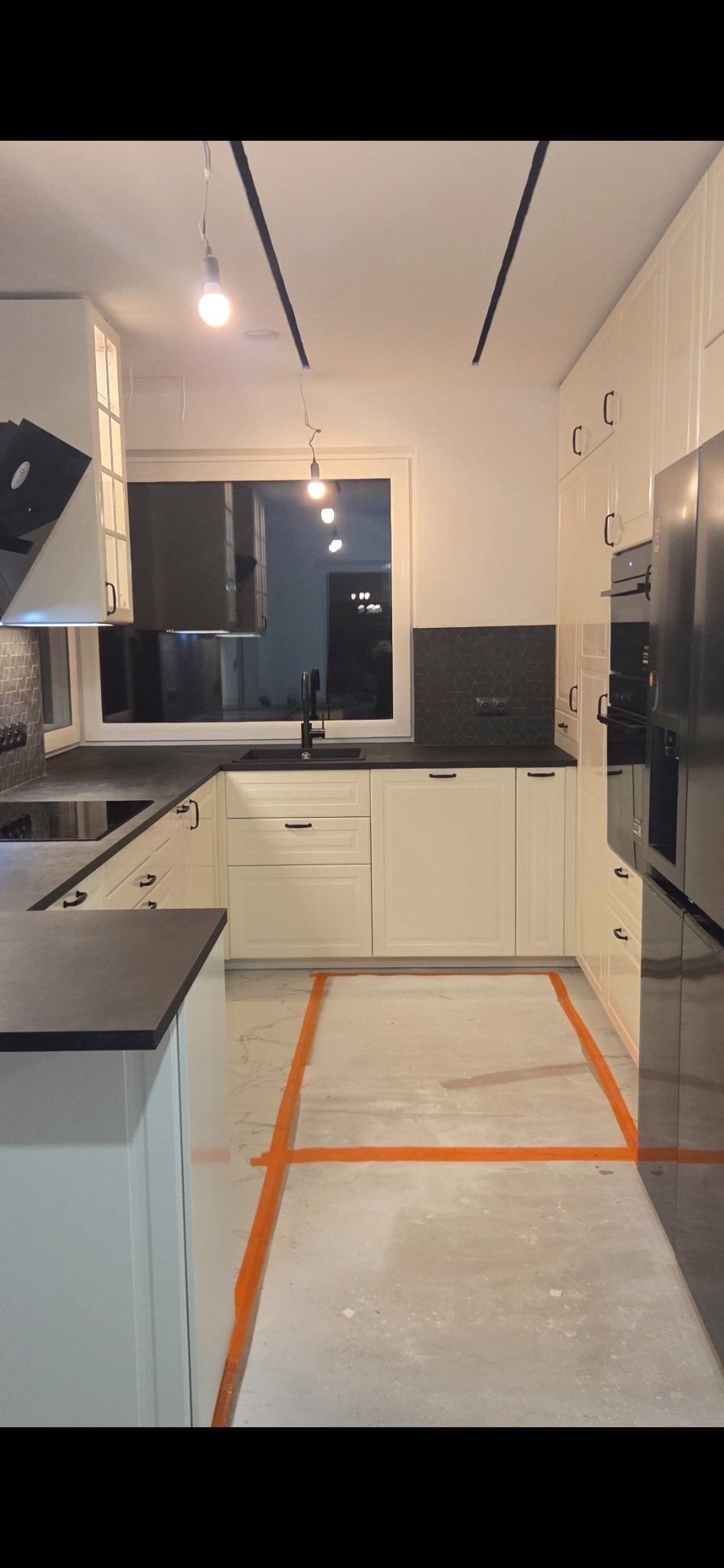Jasna kuchnia z czarnymi akcentami: blat, armatura i płytki. Podłoga przygotowana do położenia płytek, wyznaczona pomarańczową taśmą. Widok na okno i oświetlenie.