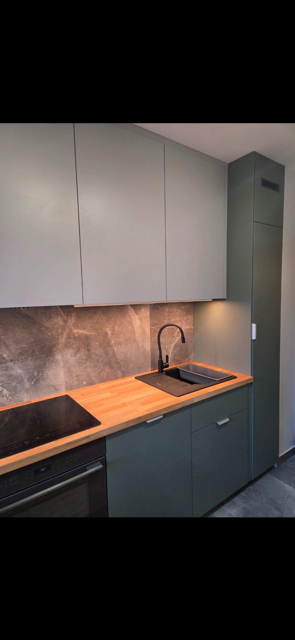 Nowoczesna kuchnia z szarymi szafkami górnymi i zielonymi dolnymi, drewnianym blatem, czarną armaturą i marmurowym panelem. Minimalistyczny design z oświetleniem podszafkowym.