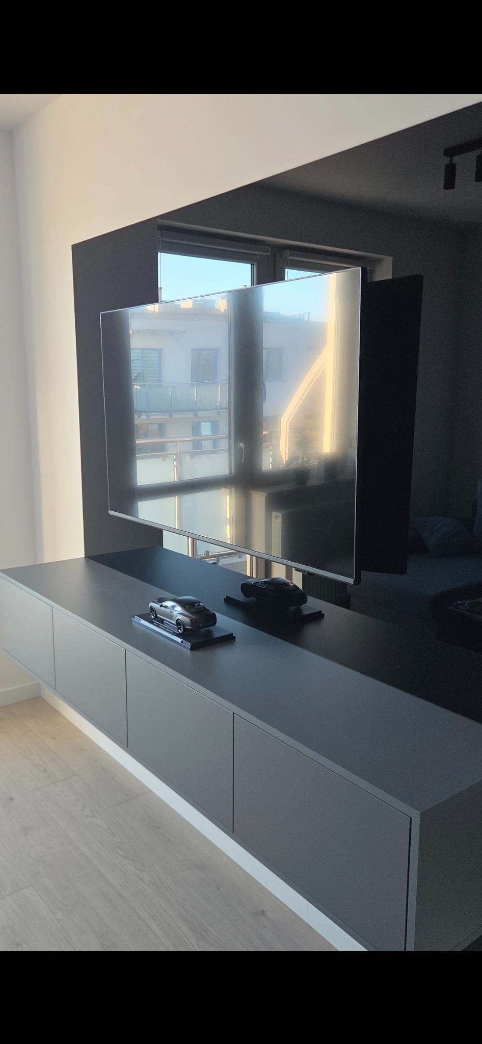 Szara, podwieszana szafka RTV z minimalistycznymi frontami, na której stoją dwa modele samochodów. W tle okno z odbiciem krajobrazu.