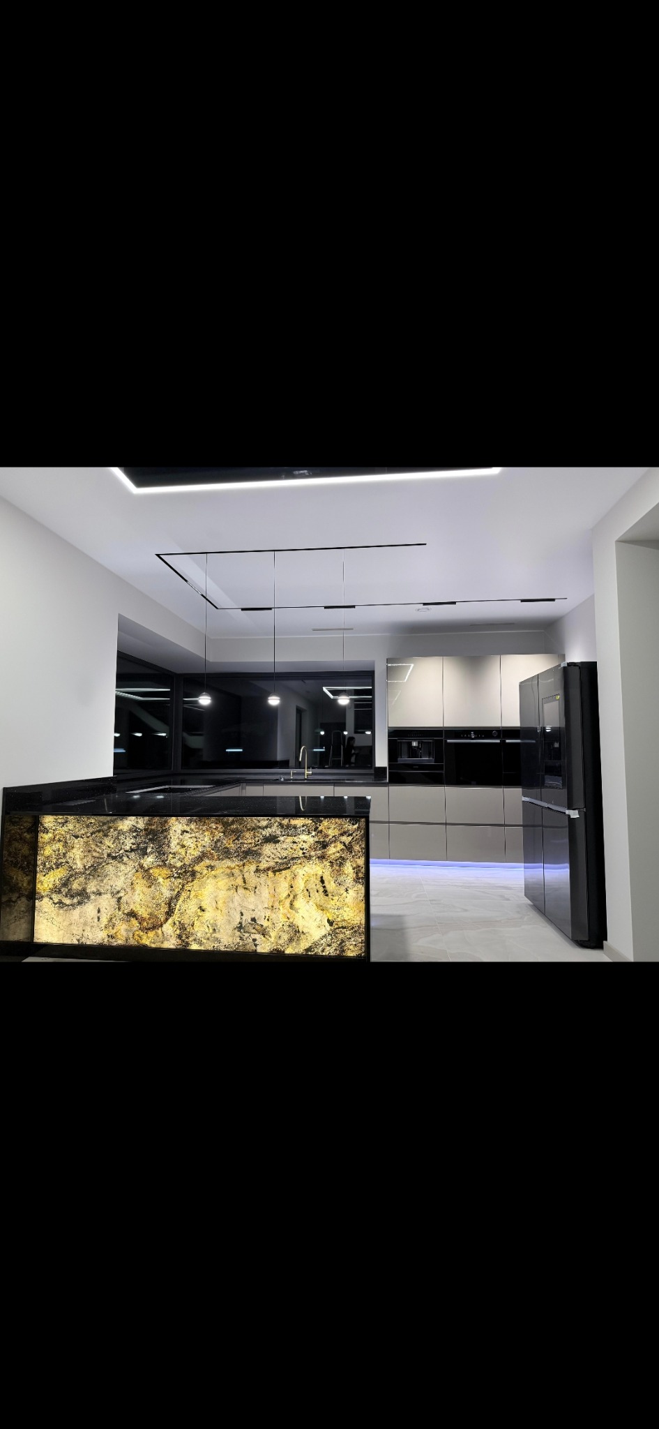 Nowoczesna kuchnia z wyspą oświetloną od dołu, fronty w odcieniach szarości i czerni, minimalistyczne oświetlenie sufitowe. Blat wyspy z czarnego kamienia.