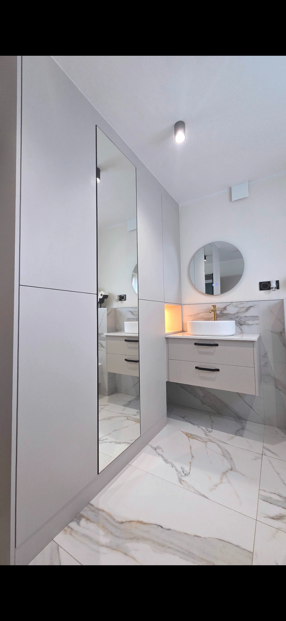 Nowoczesna łazienka z szafką pod umywalkę w odcieniach szarości, lustrem w zabudowie i marmurową podłogą. Minimalistyczny design z okrągłym lustrem i złotą armaturą.