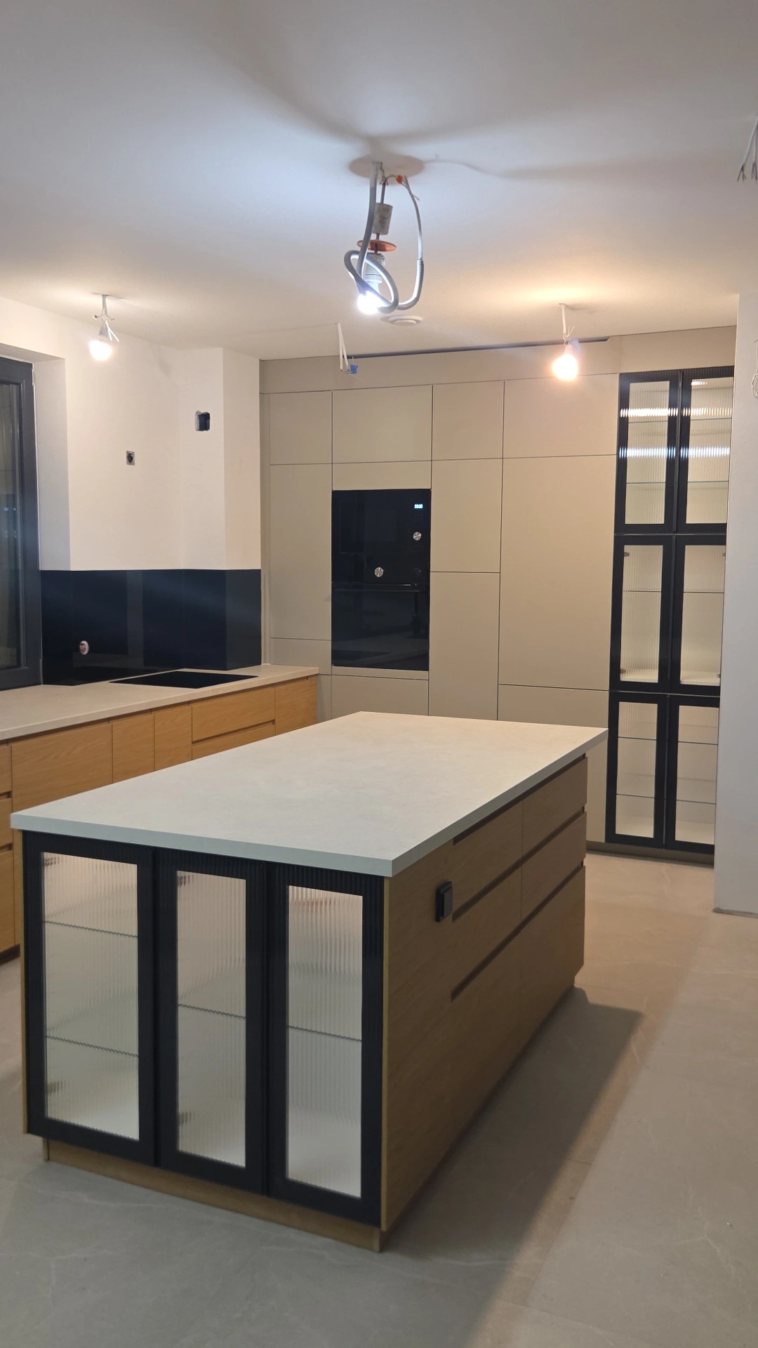 Nowoczesna kuchnia z wyspą i ryflowanymi szybami w zabudowie. Beżowe fronty szafek, czarny sprzęt AGD. Widoczne niedokończone prace wykończeniowe.
