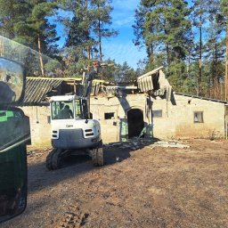 ROB-INVEST Usługi minikoparką Robert Bełdycki - Mini koparka gąsienicowa w trakcie rozbiórki starego, parterowego budynku z cegły. Widoczne uszkodzenia dachu i ścian. W tle las iglasty i błękitne niebo.