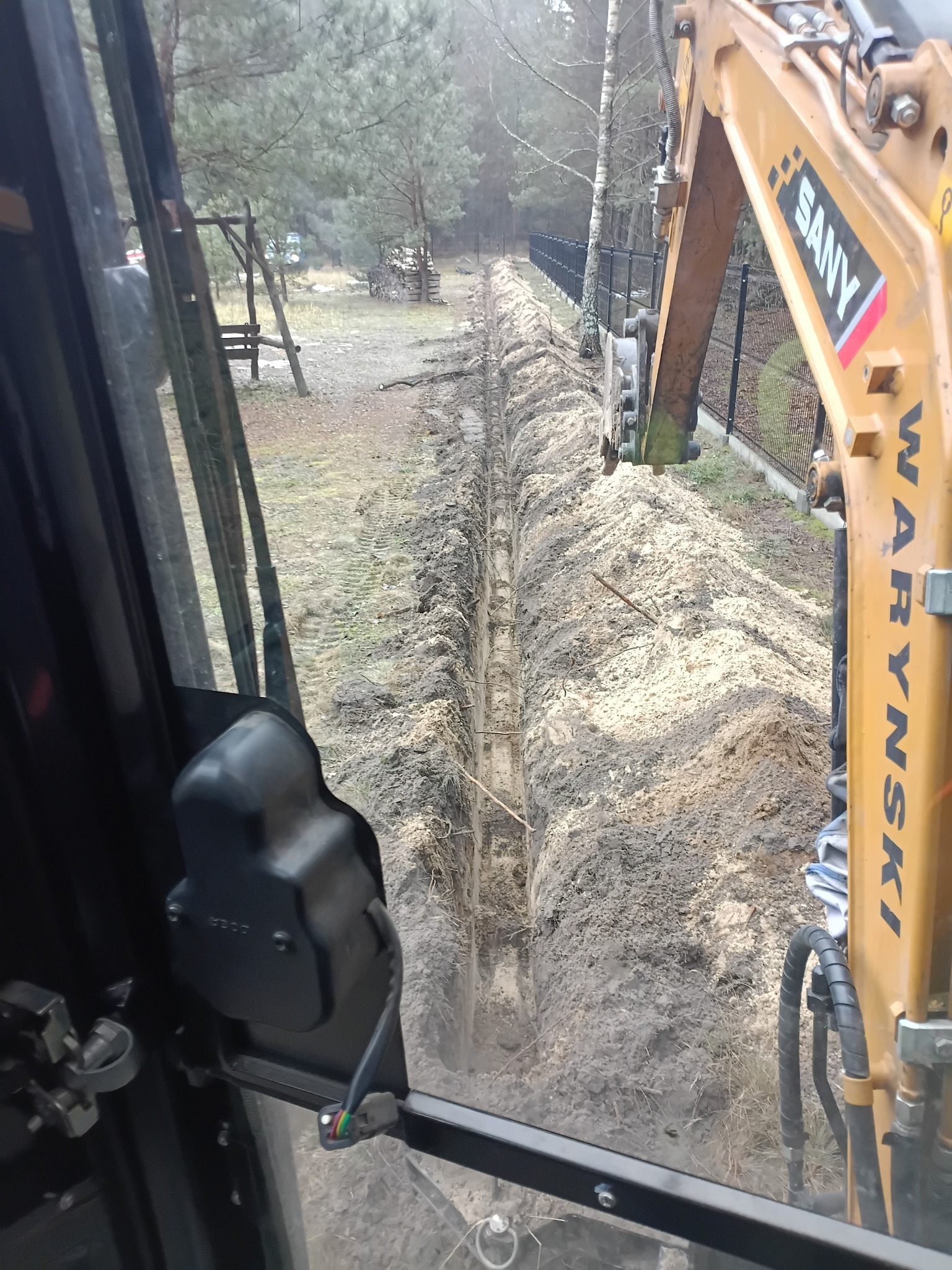 Wąski wykop w ziemi, widziany z kabiny koparki SANY WARYNSKI, obok czarne ogrodzenie panelowe. Prace ziemne w terenie zalesionym.