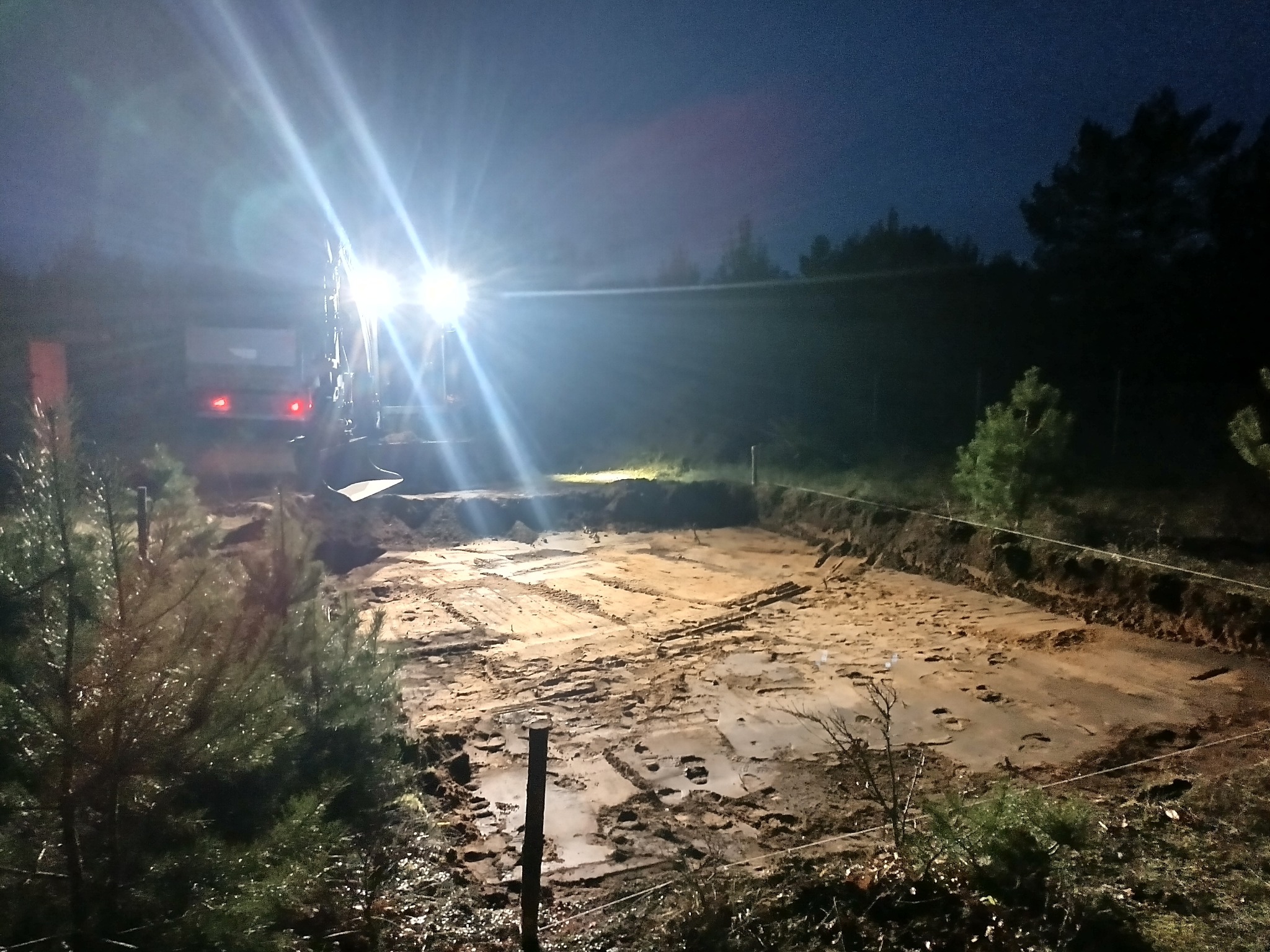 Nocny wykop pod fundamenty oświetlony reflektorami koparki. Widoczne ślady opon i wykopana ziemia. W tle ciemne drzewa i niebo.