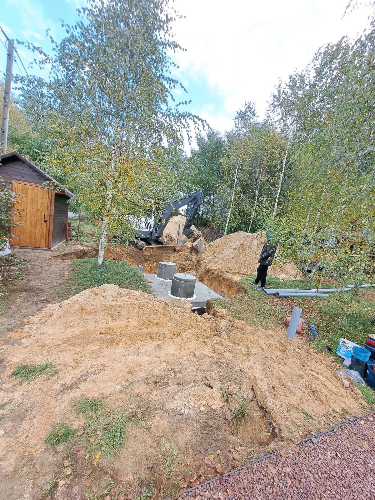 Wykop pod szambo z betonowymi kręgami, częściowo zasypany ziemią. Koparka w tle, obok rury i narzędzia. Widok z góry na wykop w ogrodzie.