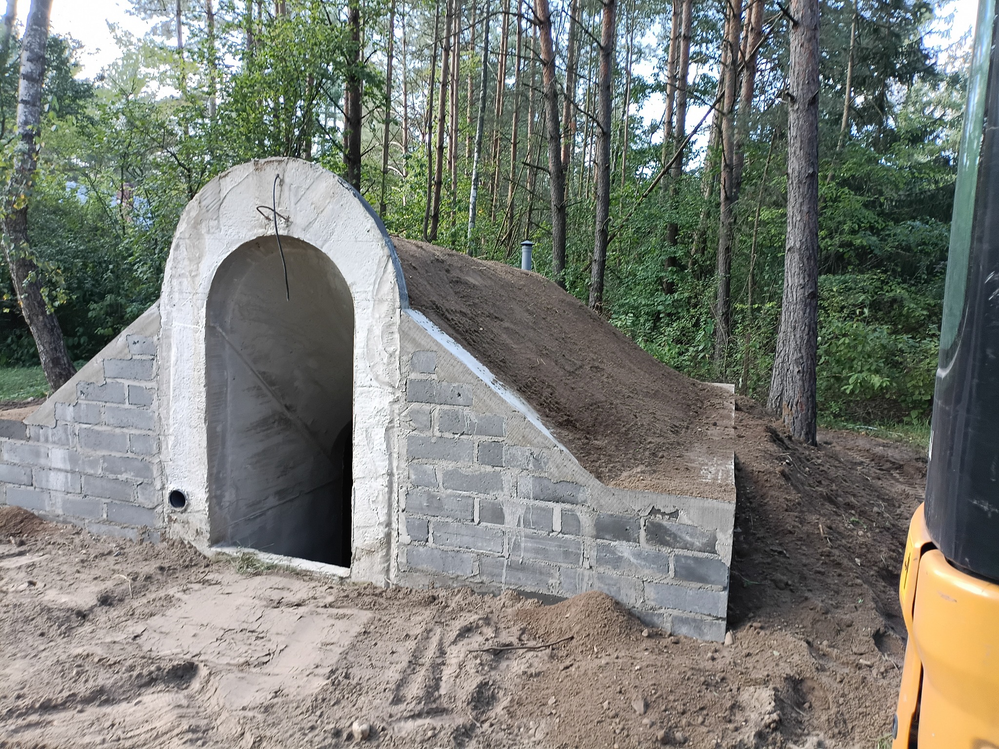 Ziemianka z wejściem z szarej cegły, częściowo przysypana ziemią, na tle lasu. Widoczny fragment maszyny budowlanej. Nietypowa konstrukcja w naturalnym otoczeniu.