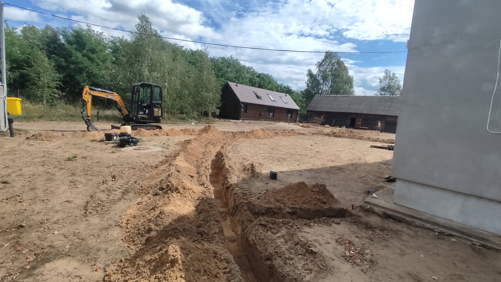Żółta koparka przy wykopie pod instalacje na piaszczystym terenie budowy, w tle budynki i drzewa. Wykopane rowy widoczne na pierwszym planie.