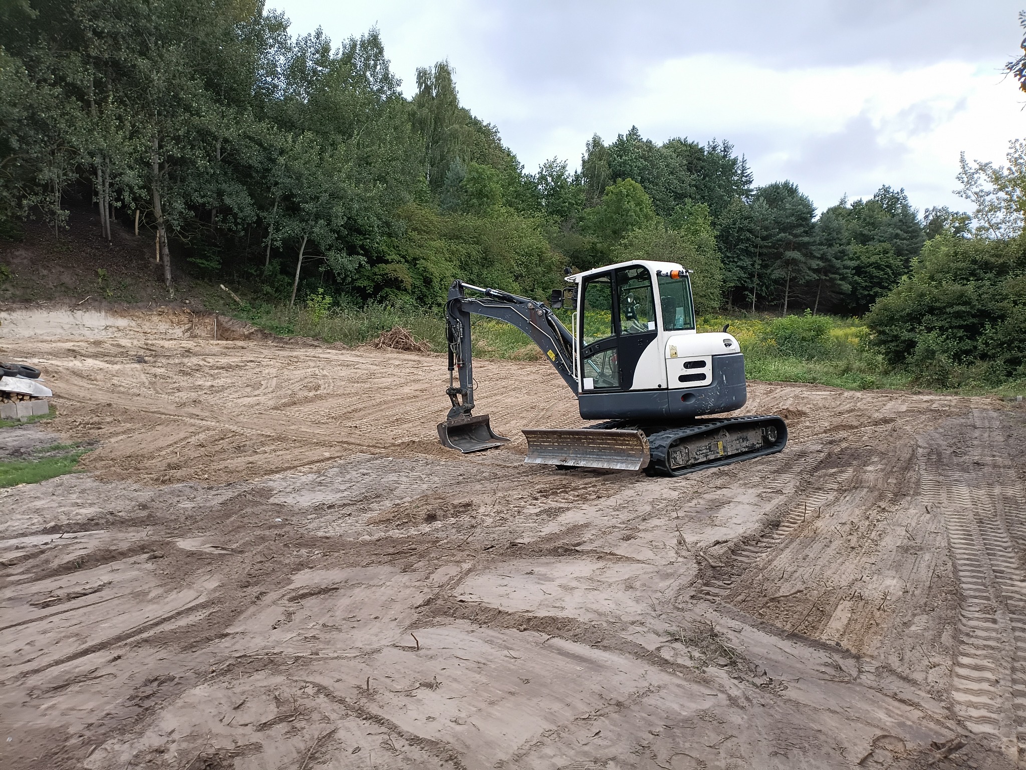 Mała koparka na placu budowy z widocznymi śladami opon na piasku i drzewami w tle, sugerująca prace ziemne w toku.