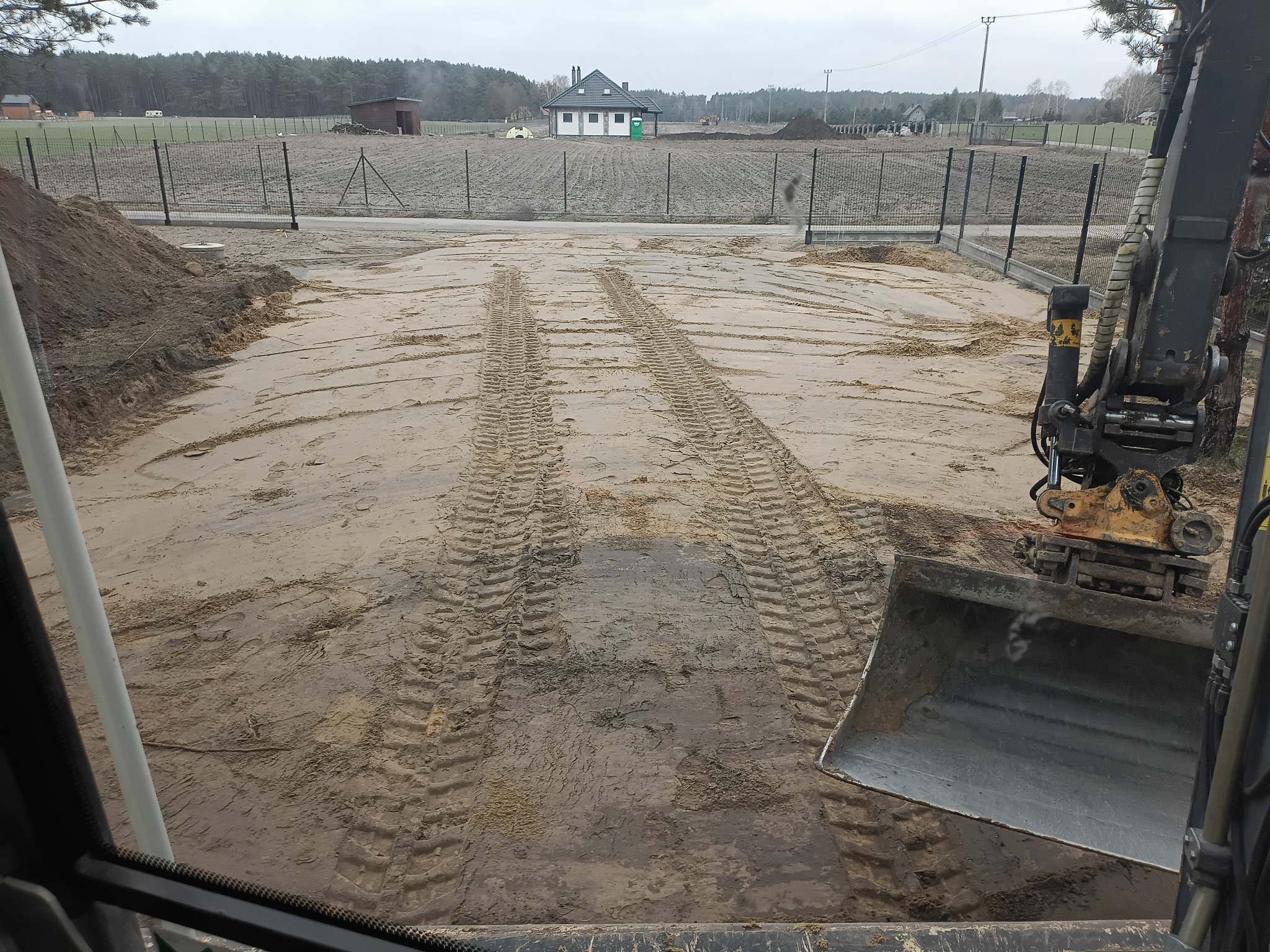 Ślady opon koparki na wyrównanym, piaszczystym terenie budowy z widocznym ogrodzeniem i domem w tle. Ujęcie z kabiny maszyny.