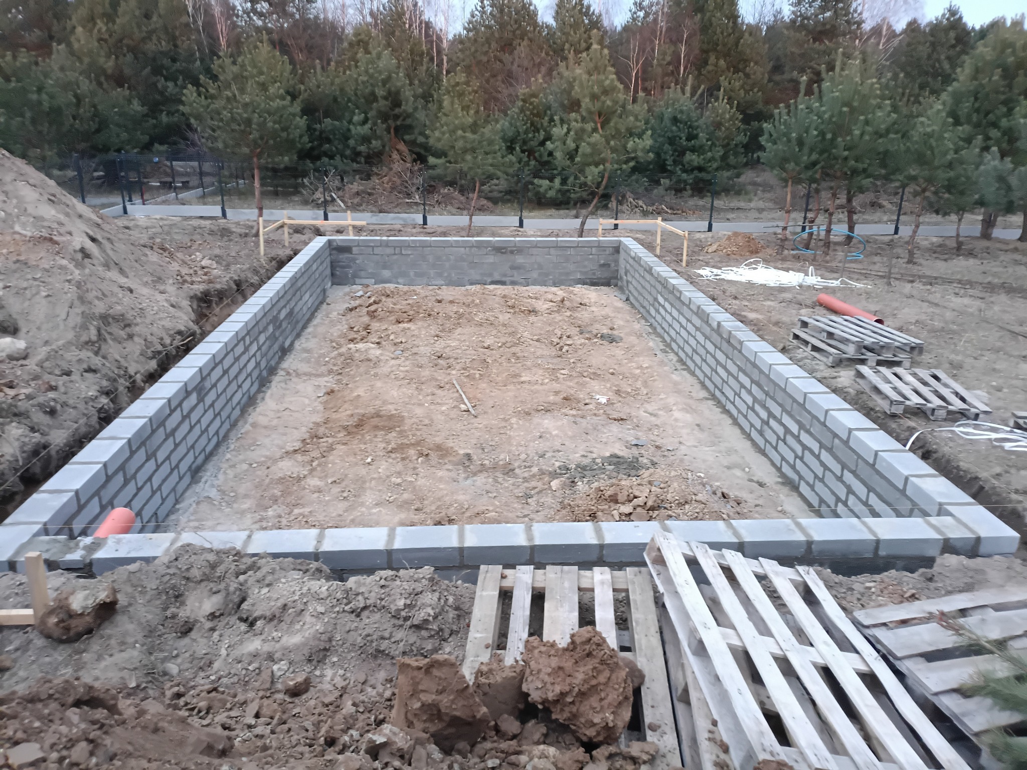 Wykop pod fundamenty z częściowo wzniesionymi ścianami z bloczków betonowych. Widoczne palety, rury i wykopana ziemia. W tle drzewa i ogrodzenie.